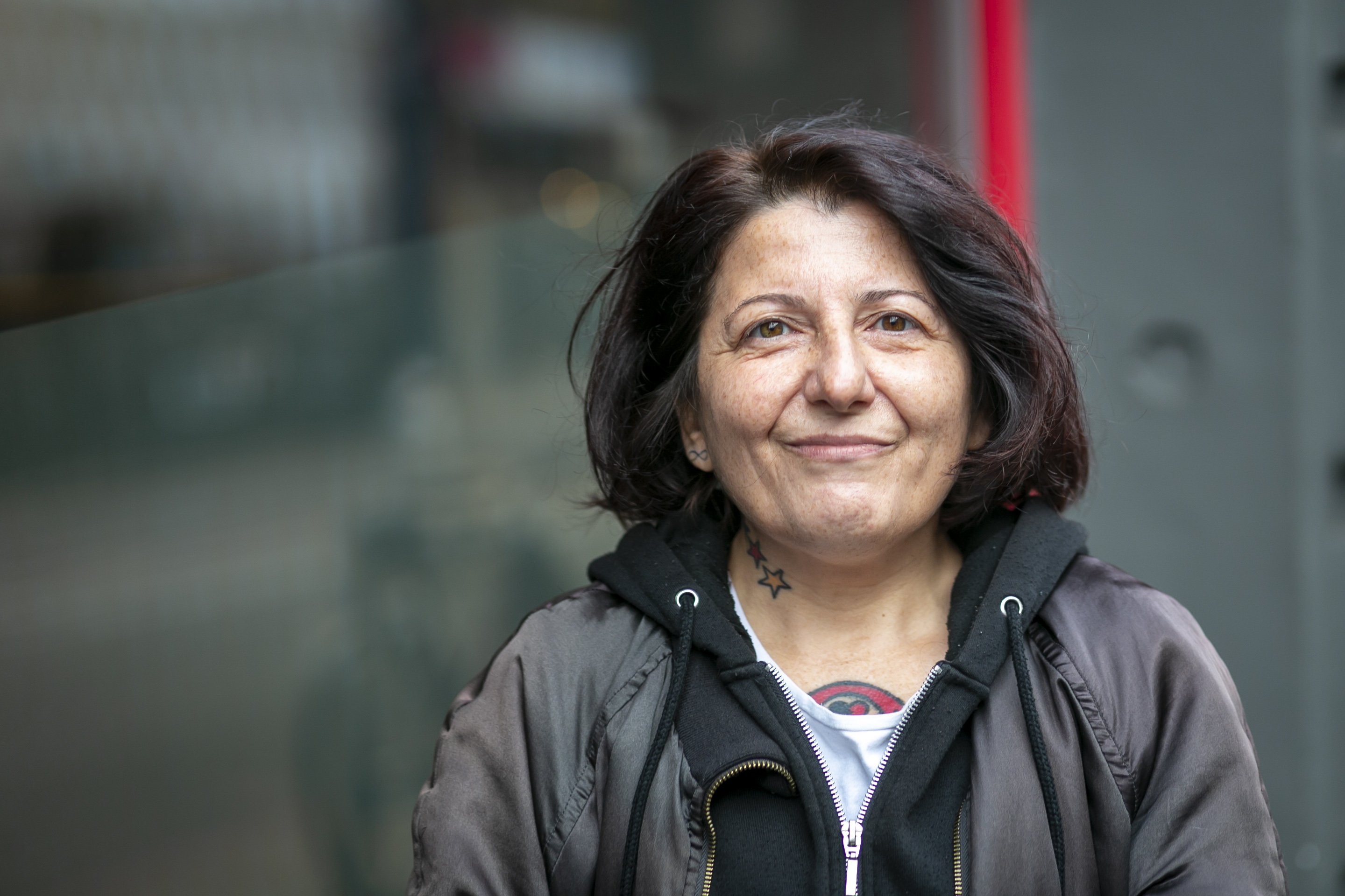 A smiling woman with dark hair wearing a dark jacket and hoody looks directly at the camera