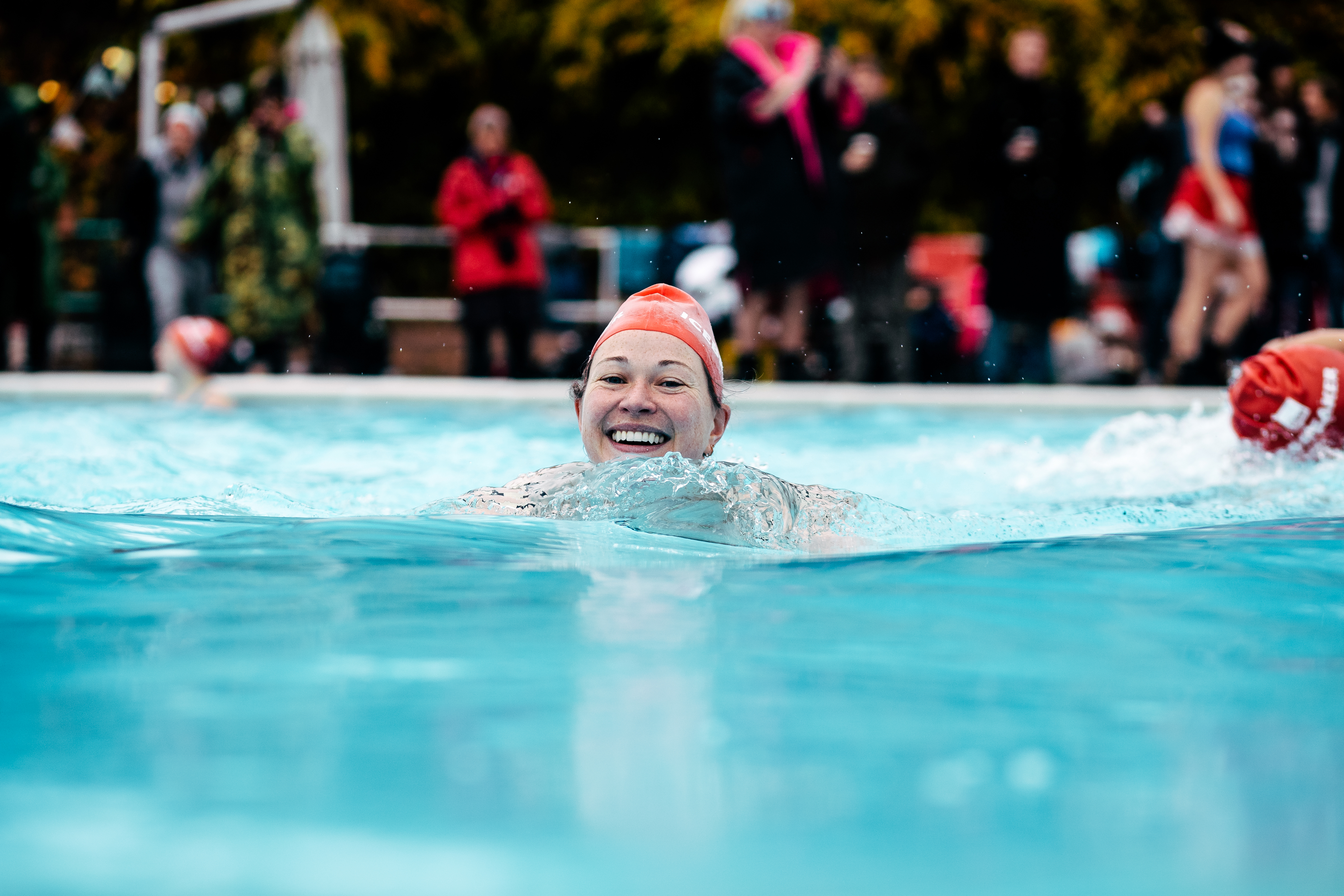 A woman wearing a red Crisis branded swimming cap smiles in the lido.