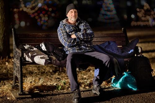 Nick is sitting on a bench at nighttime. On the bench and floor around him are his belongings in various bags.