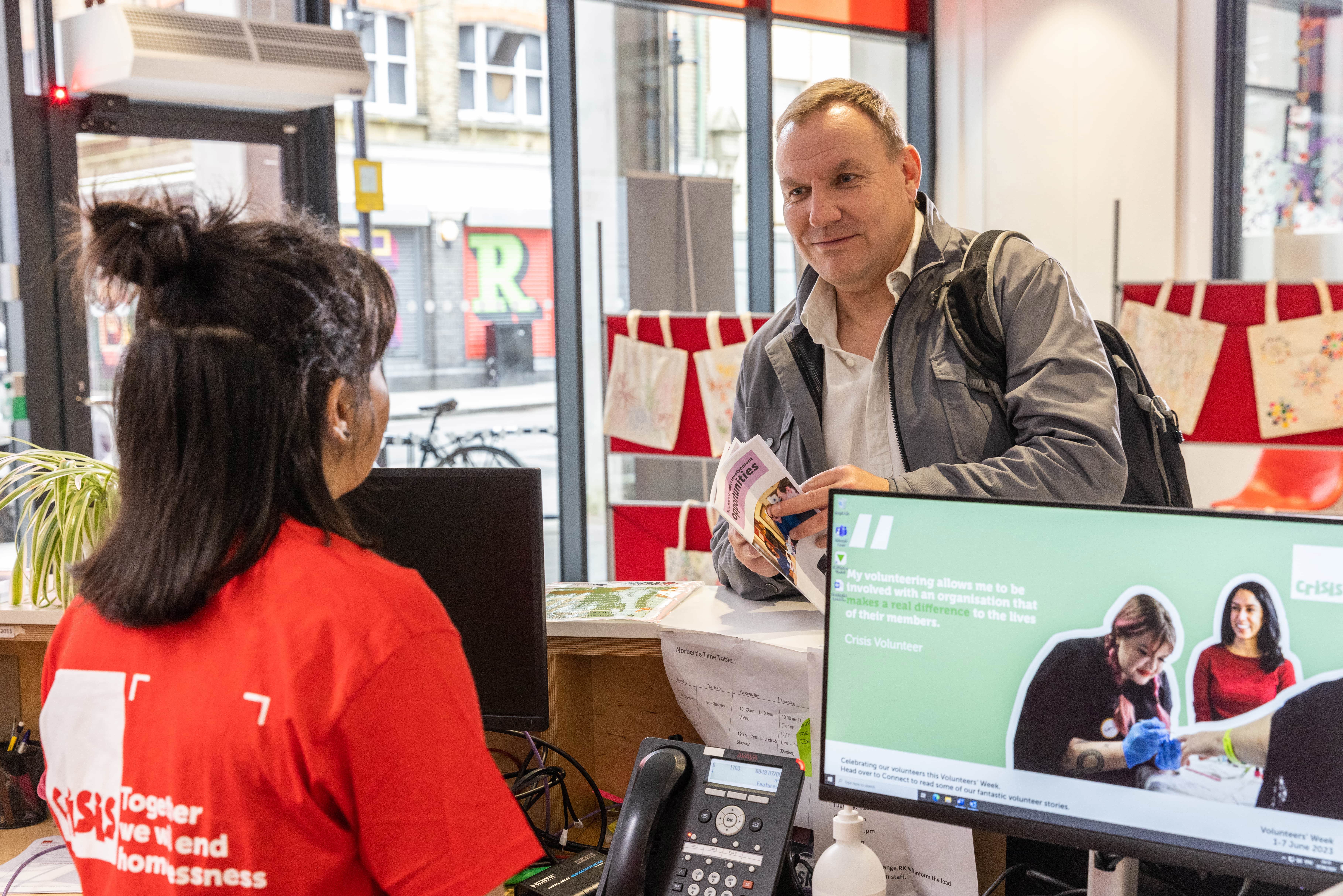 A volunteer, wearing a red Crisis t-shirt, welcomes a man to a Crisis Skylight centre at a reception desk. 