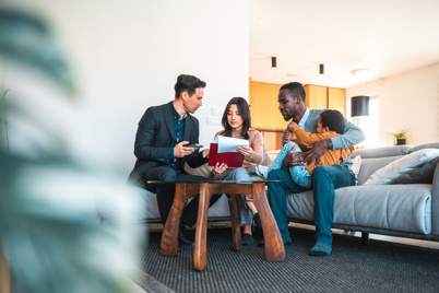 Two adults and their child are sat around a coffee table in the living area of a flat talking with a man wearing a suit. The man in the suit is showing them documents. The atmosphere is relaxed and comfortable.