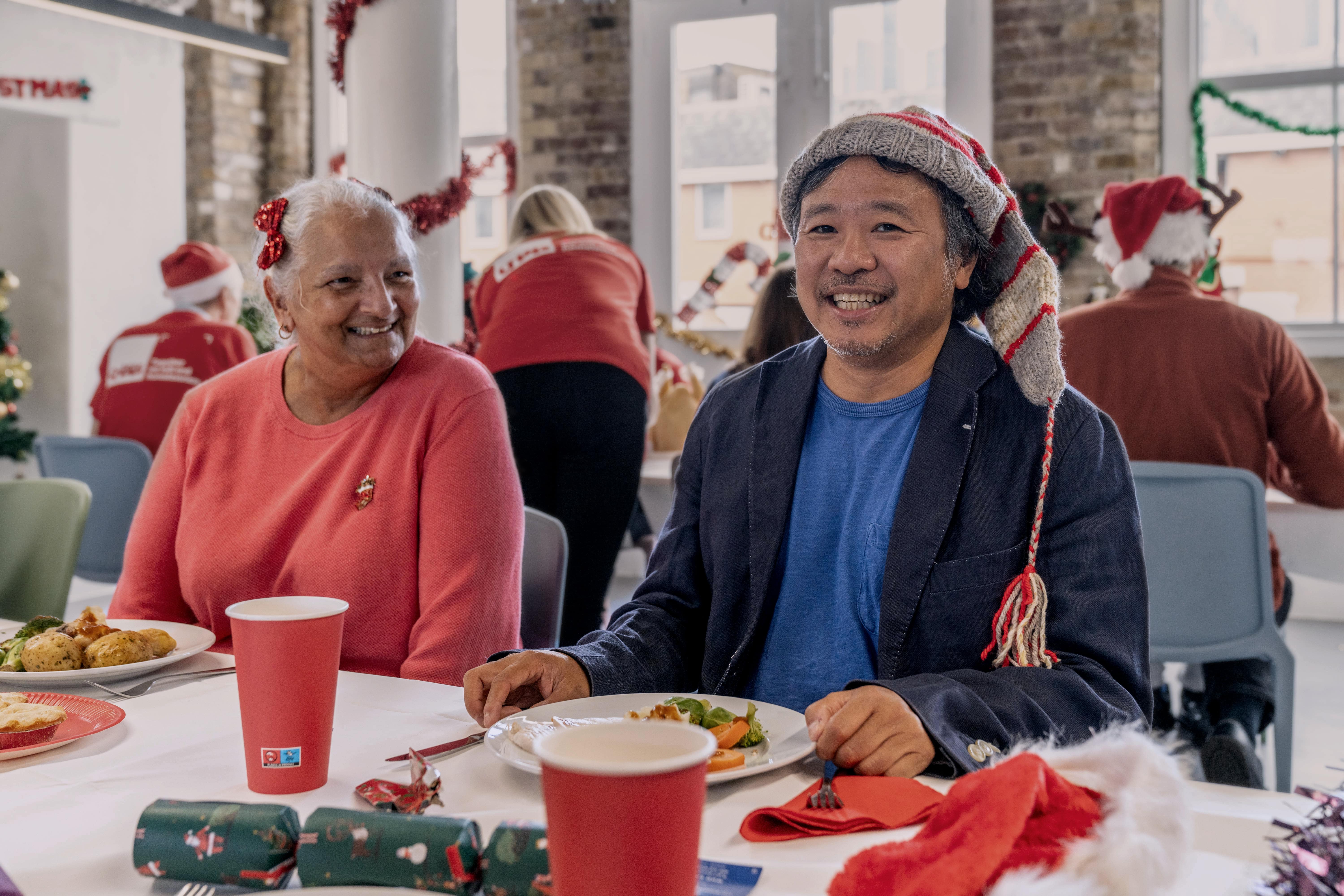 A man and a woman sit at a Christmas dinner table, smiling at the camera. 