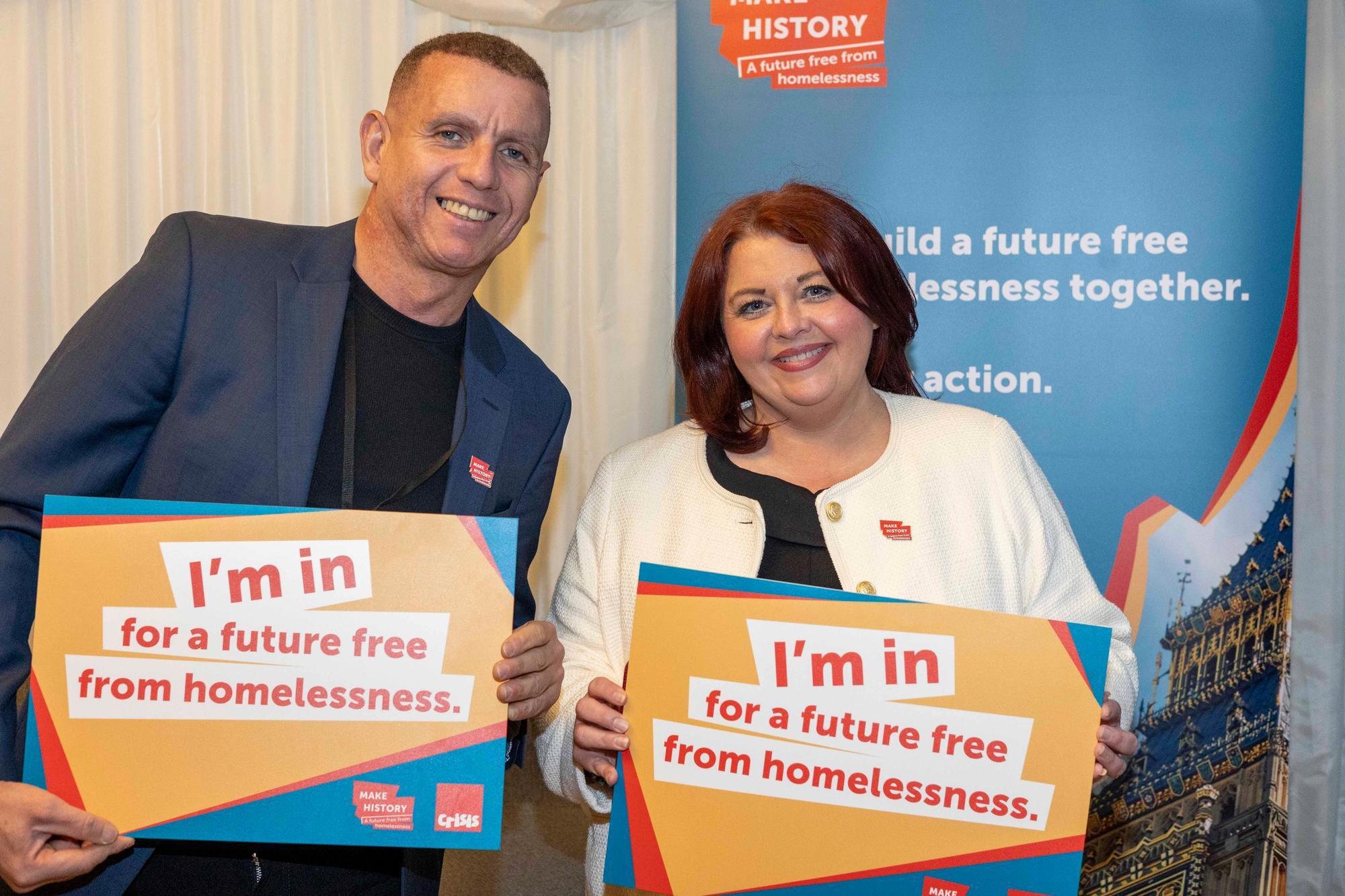 A man in his 40s wearing a blue suit stands with a women in her 50s wearing a white jacket. They're both holding signs that say 'I'm in for a future free from homelessnes'.