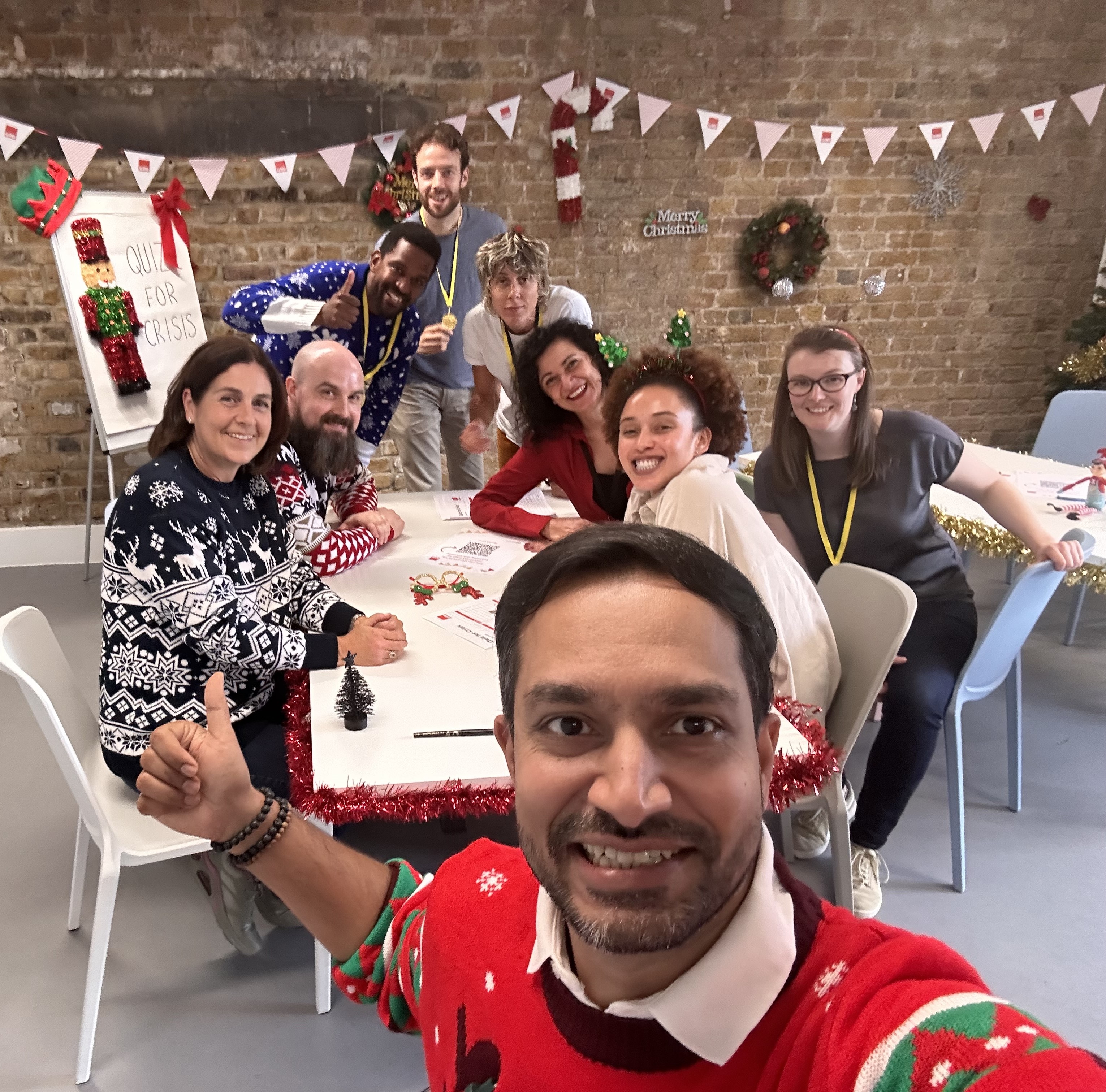 A man wearing a red Christmas jumper takes a selfie at the front of a group of people around a table.  There are festive decorations and everyone is smiling at the camera.