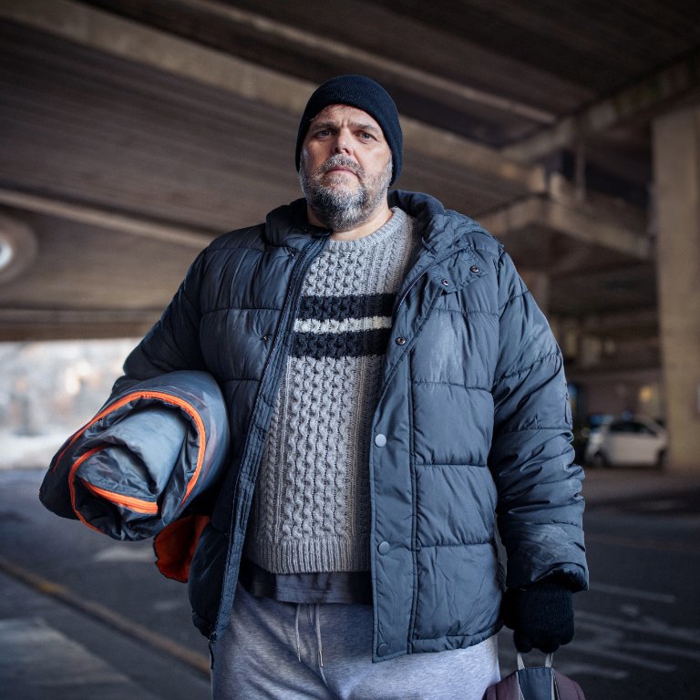 Gaz stands under a bridge holding a sleeping bag, dressed for cold weather, looking directly at the camera.