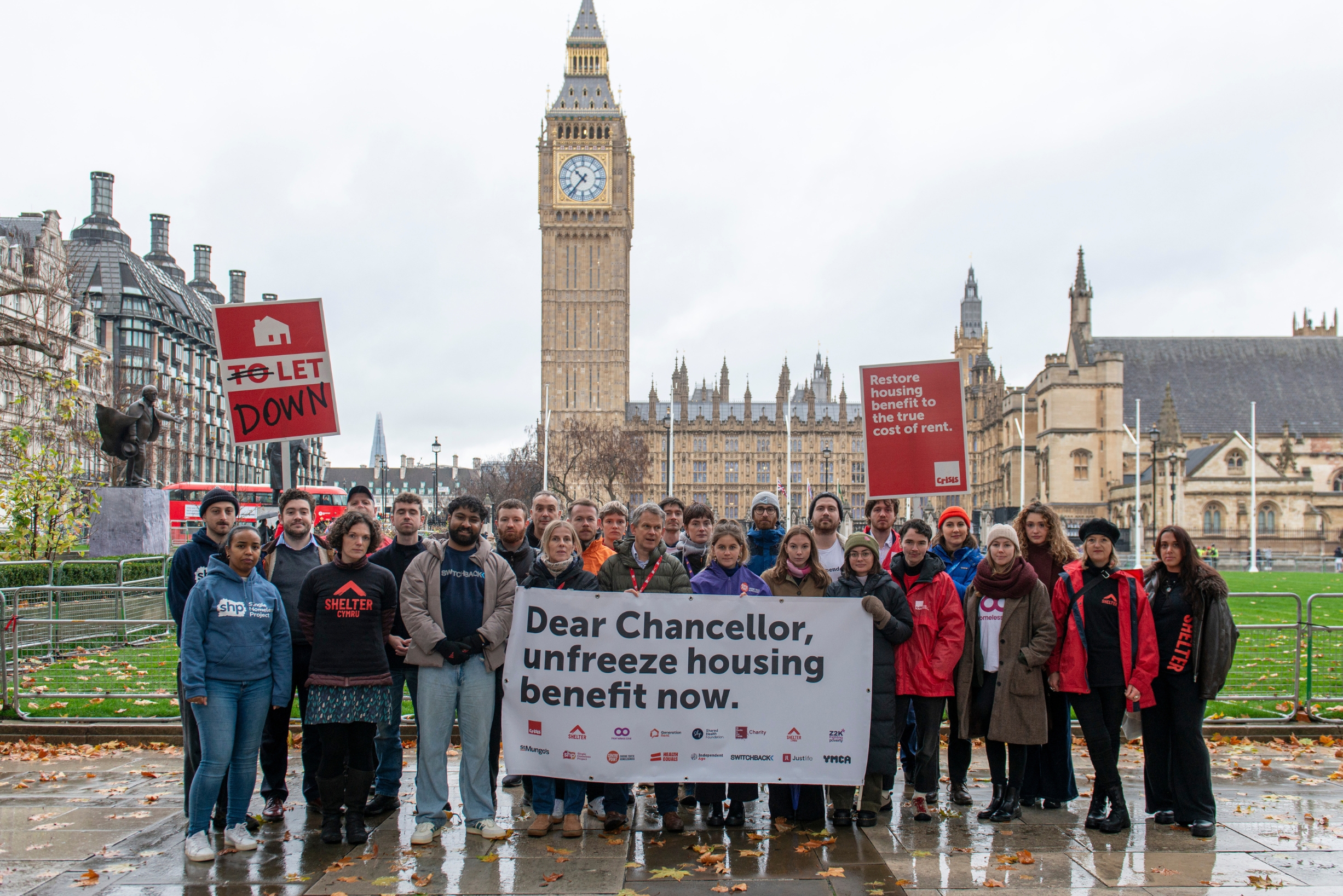 A group of thirty people dressed in winter clothes standing in Parliament Square holding a banner that says 'Dear Chancellor: unfreeze housing benefit now.'
