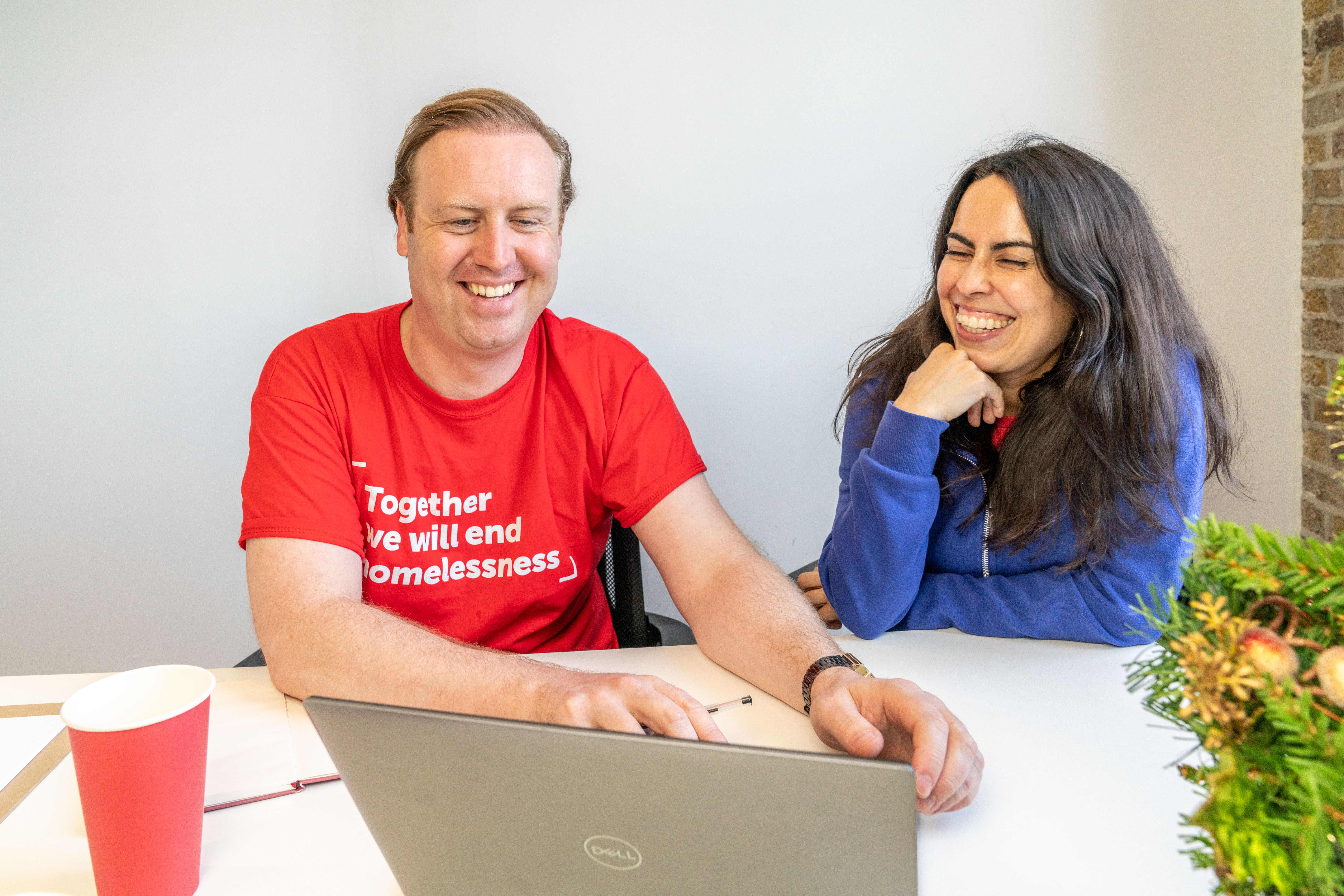 A man wearing a red Crisis branded t-shirt and a woman look at a laptop together. They are both smiling. 