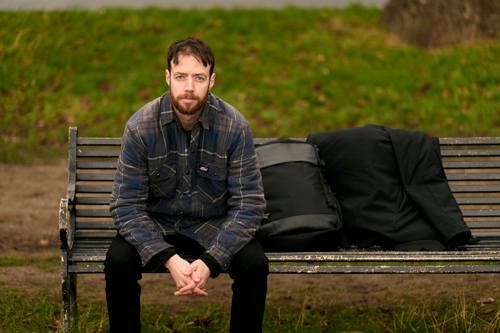 Bradley sits on a park bench looking at the camera. His rucksack and coat are placed next to him.