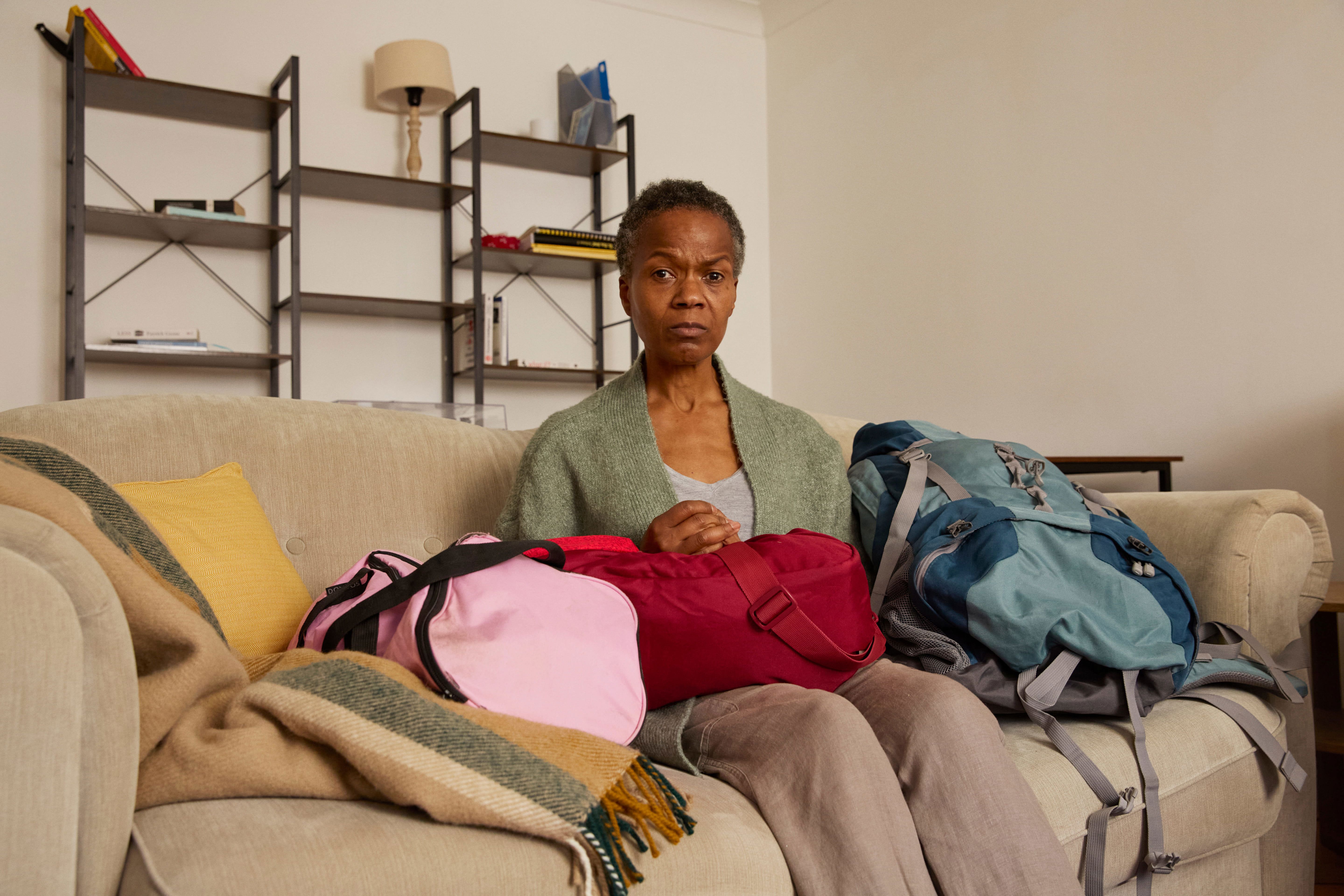 Gabrielle is sitting on a sofa in a living room with sparse decoration. Around her, on the sofa and the floor, are various bags. 