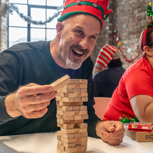 A Crisis member plays Jenga while wearing a Christmas elf hat. He is smiling and laughing. 