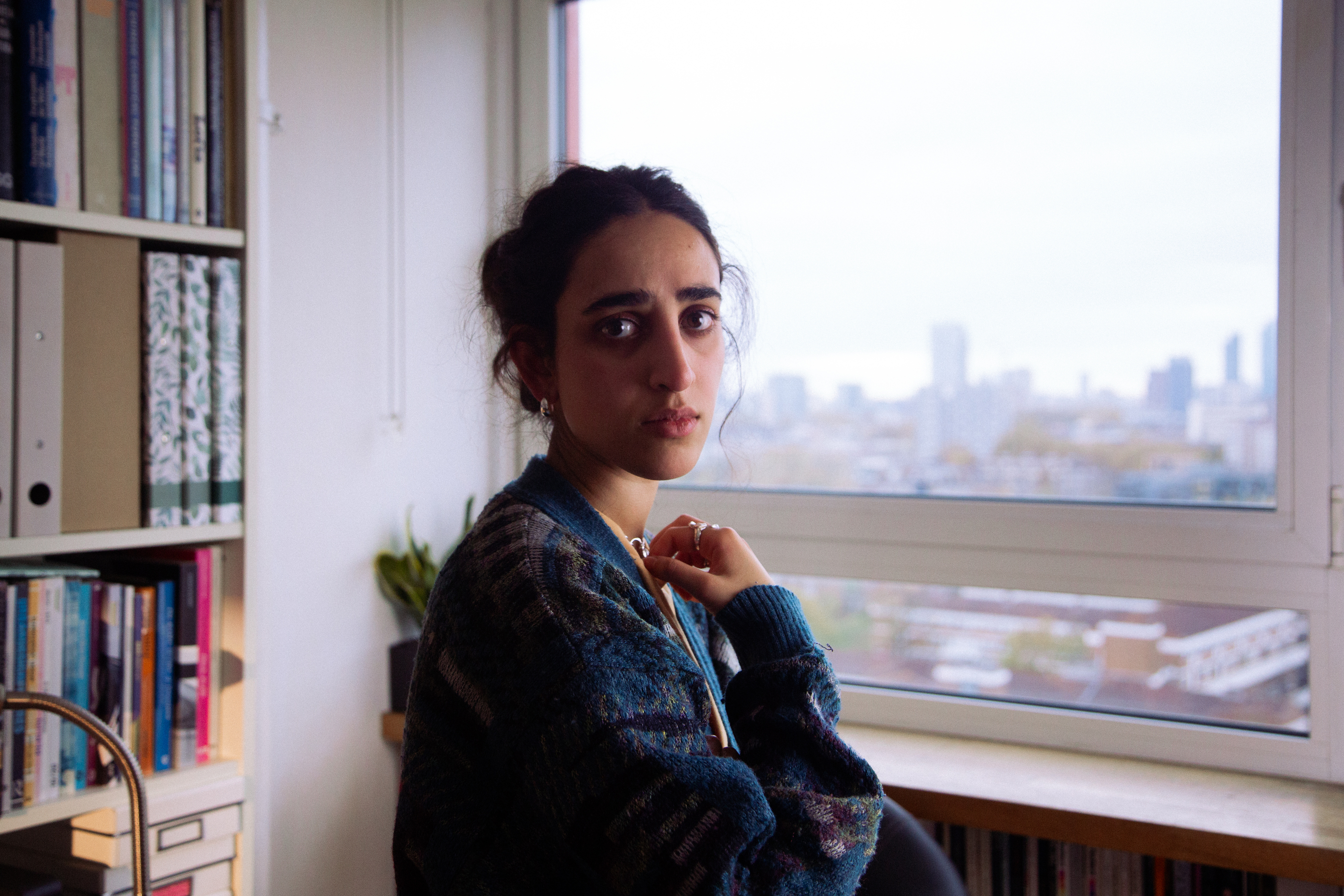 A  young woman standing by a window in a tower block of flats, looking directly at the camera.