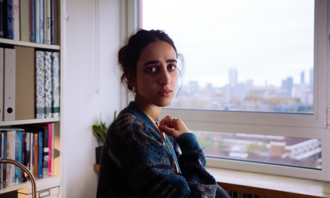 Home A young woman standing by a window in a tower block of flats, looking directly at the camera.