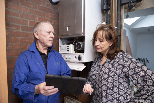 A man in a blue boiler suit shows a woman something on a tablet device. They are standing in front of a boiler which has the front panel open.