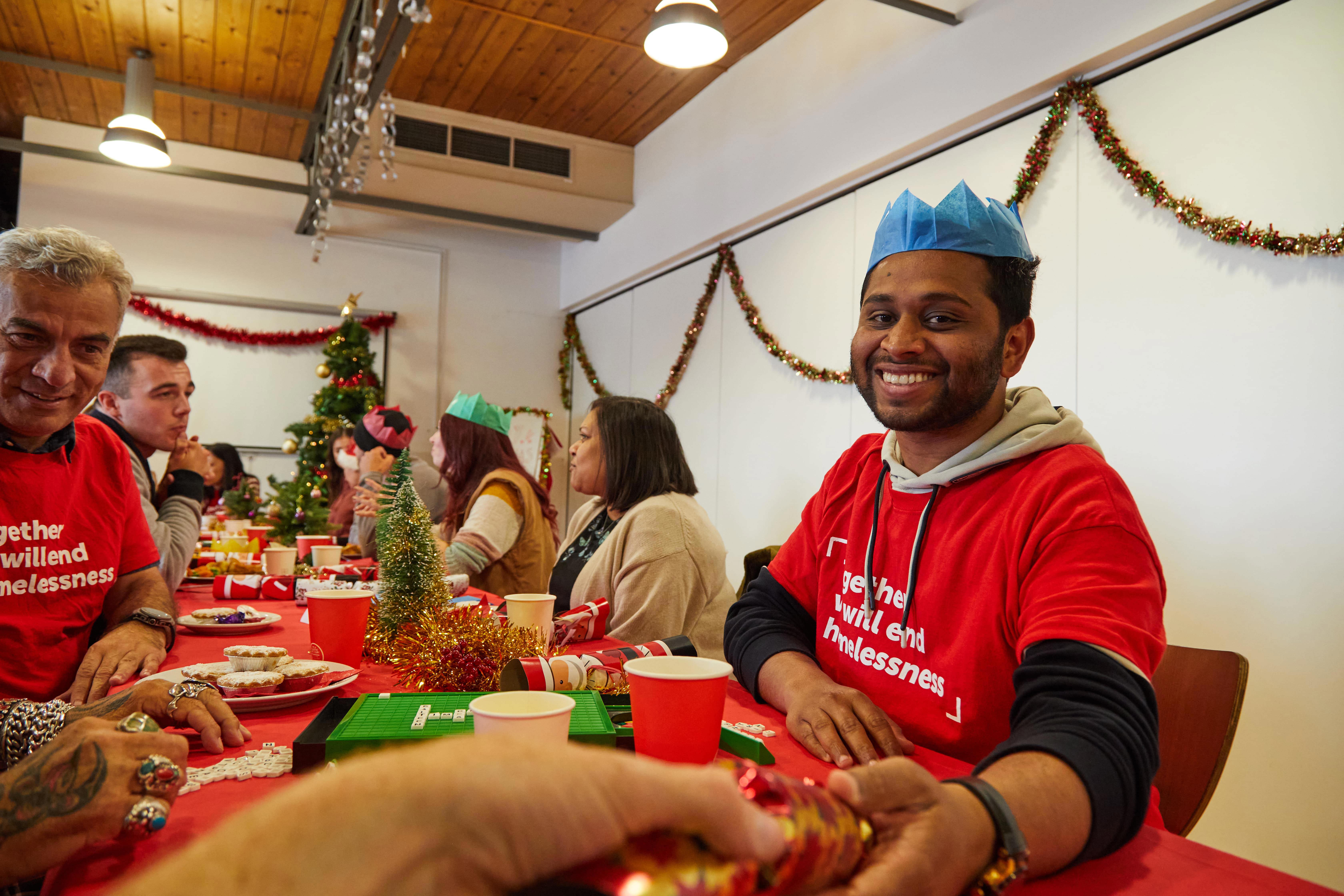 Crisis volunteers and guests eat Christmas dinner and pull crackers at a long table with Christmas decorations. 