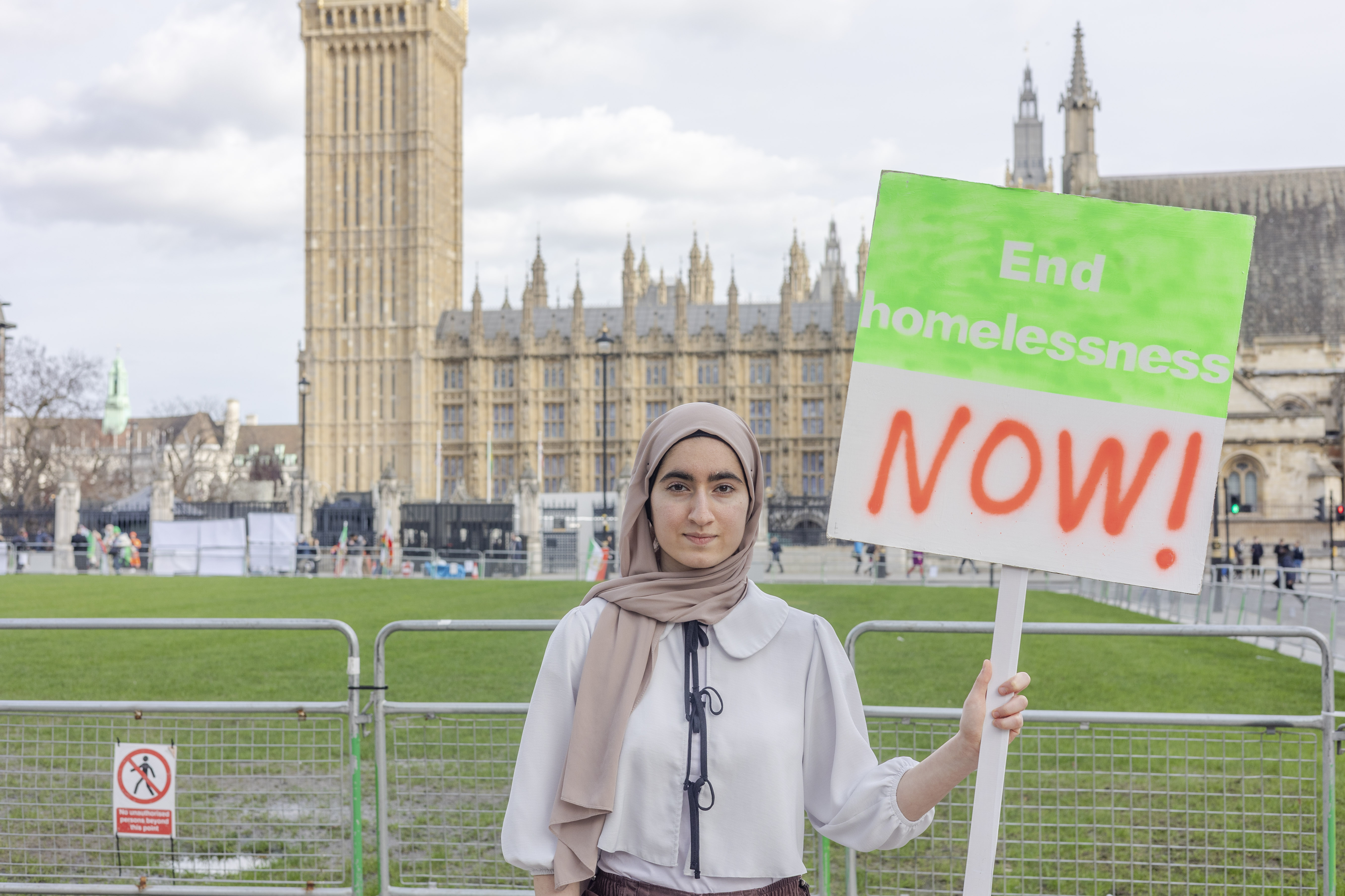 A woman wearing a headscarf stands outside Parliament holding a placard that says 'End homelessness NOW!' 